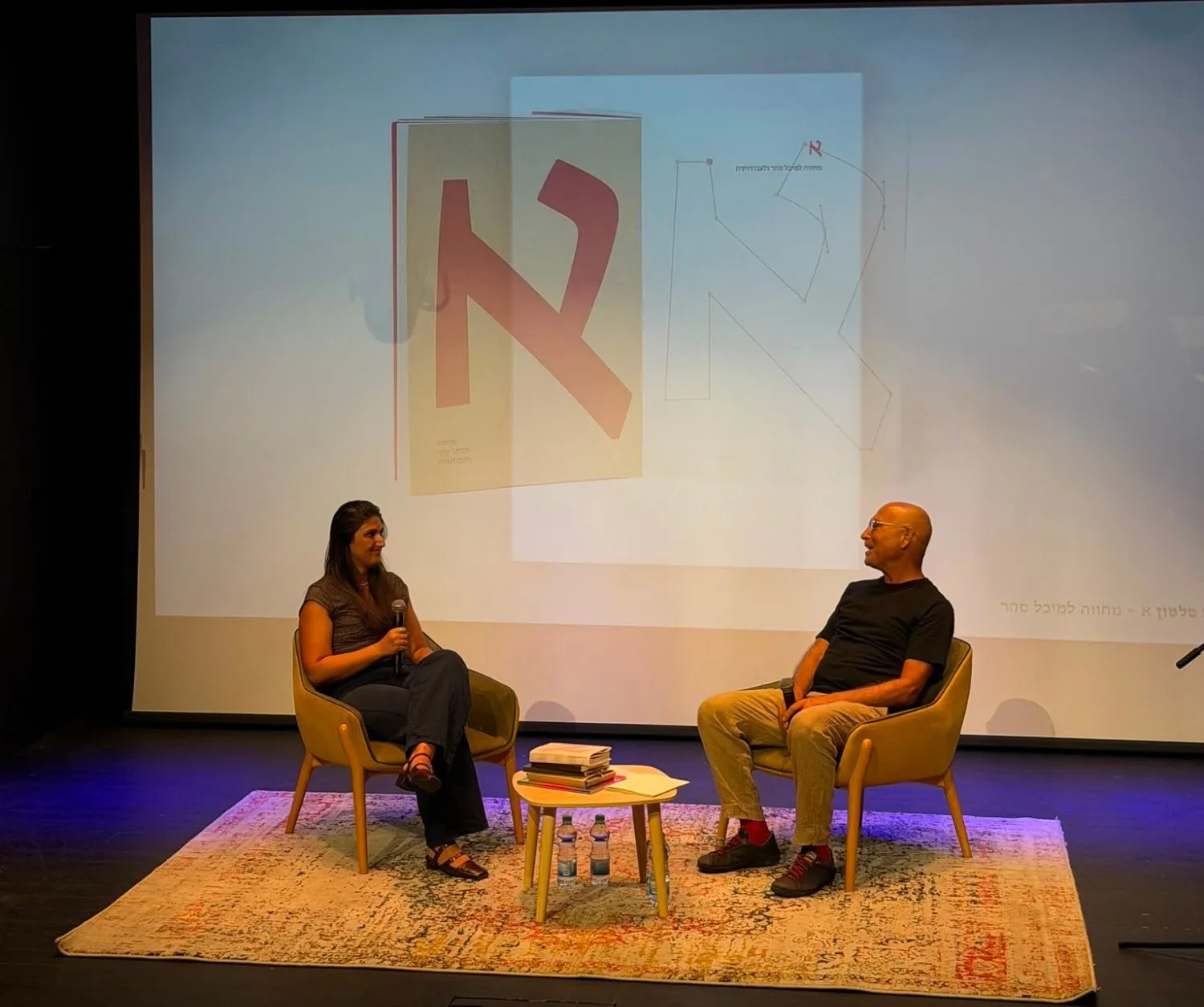 Dana on stage at the closing event at Beit Ariela, in conversation from two armchairs, the catalogue's cover and jacket projected on the screen behind them