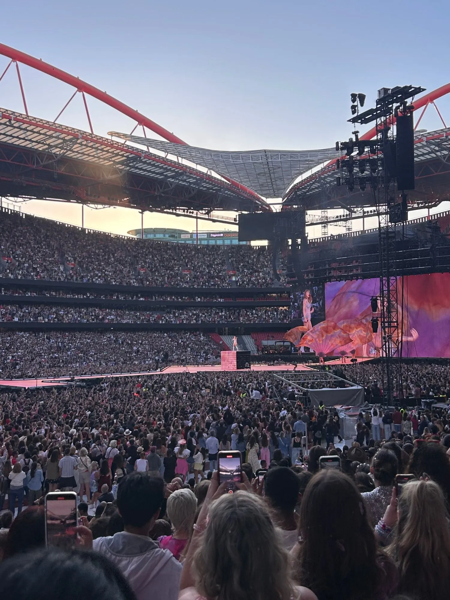 A stadium concert at dusk, the crowd filling the pitch and the stands, the performer small on a lit stage