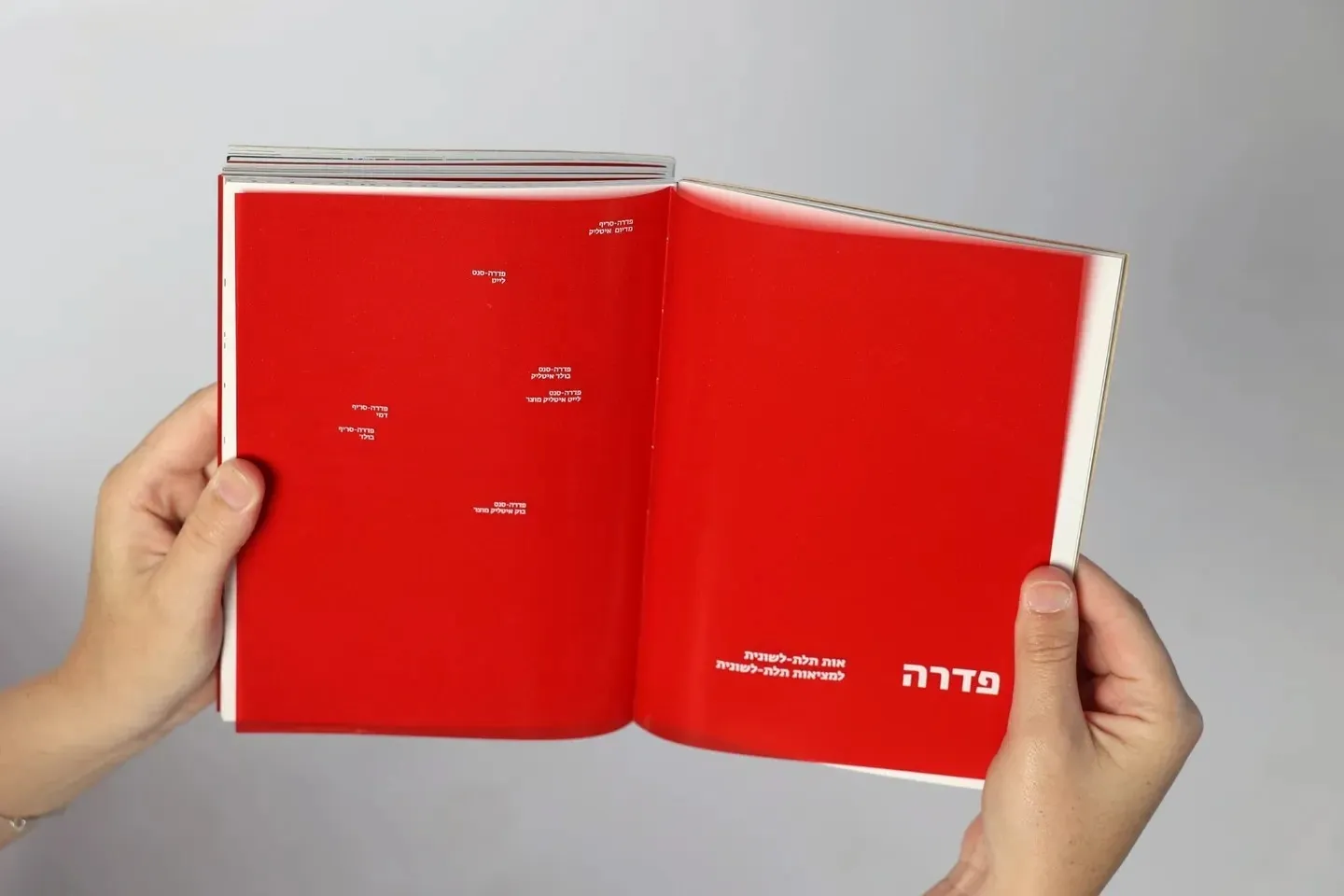 Two hands holding the catalogue open at the Fedra chapter: both pages red under the film, the running text gone, only the small white names of the weights and the title Fedra showing