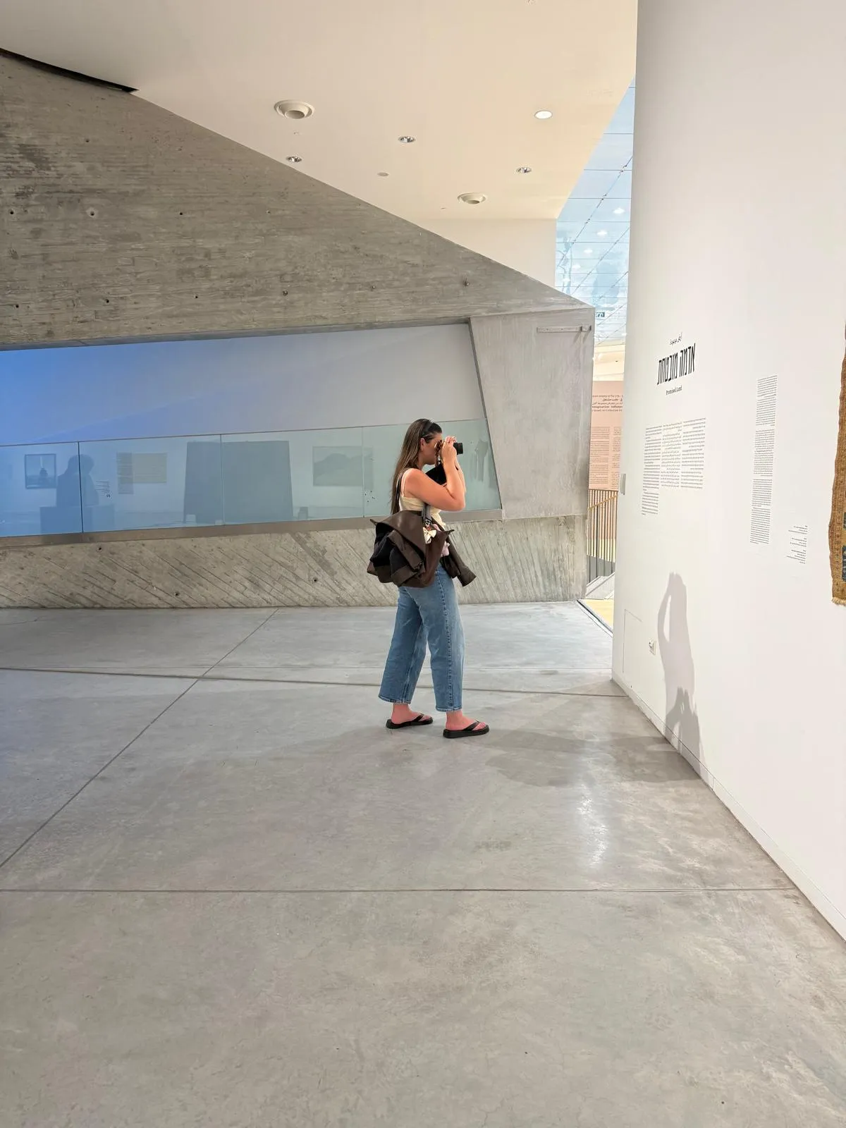 Dana photographing an exhibition wall at the Tel Aviv Museum of Art, the Hebrew title and text set in a typeface on the white wall beside her