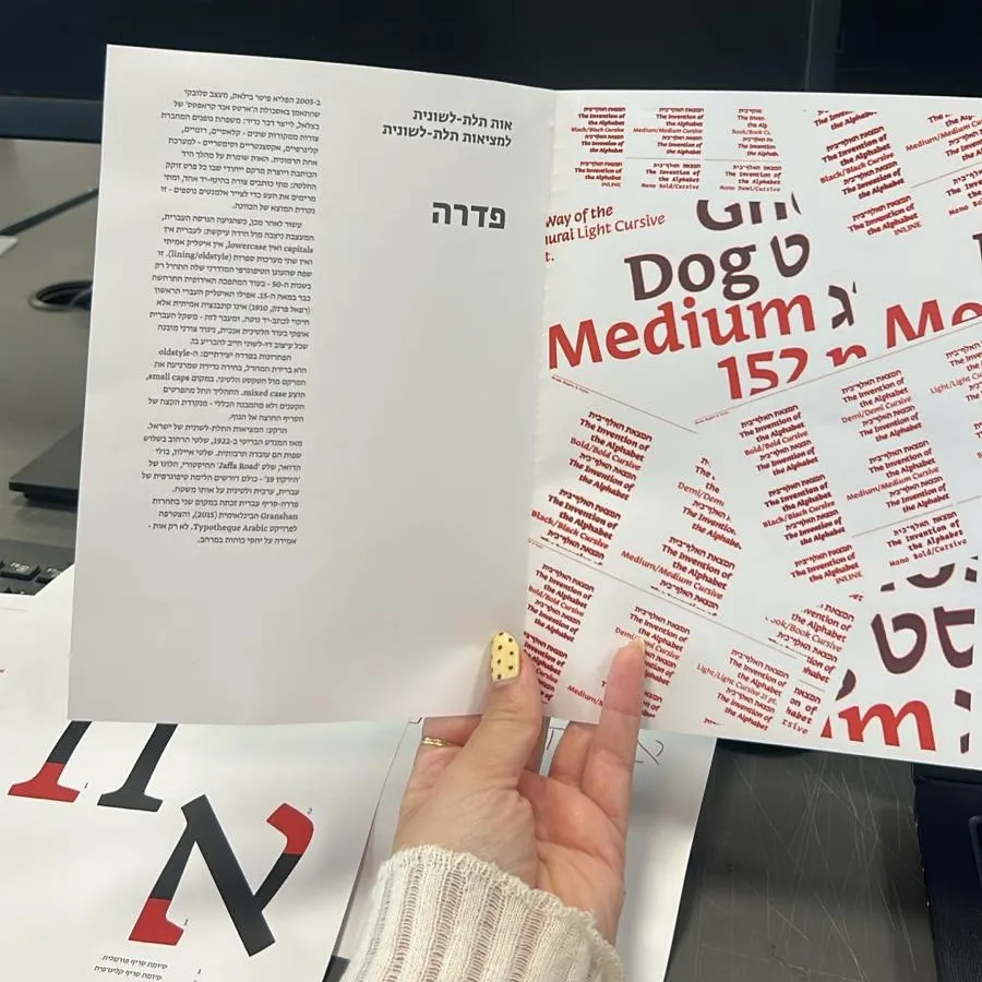 A hand holding an early print of the Fedra chapter opener, its right page covered in red and black specimen words, and on the table a sheet with a large Aleph in red and black